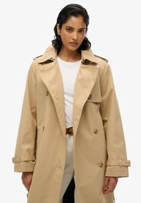 Beżowy płaszcz trencherowy z podwójnym zapięciem, dużymi klapami i mankietami z paskiem. Gładka faktura, długość do kolan, z bocznymi kieszeniami.