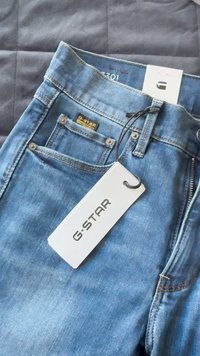 Calças de ganga azul com um design clássico de cinco bolsos, apresentando rebites metálicos, uma etiqueta preta da G-Star e um rótulo de tamanho visível no interior.