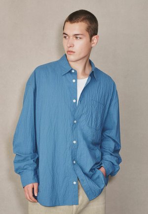 Junger Mann mit kurzen Haaren, der ein locker sitzendes, zerknittertes blaues Hemd über einem weißen Oberteil trägt und zur Seite schaut, während eine Hand in der Tasche steckt.