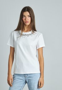 Camiseta de algodón blanca con mangas cortas y adornos decorativos alrededor del escote, combinada con jeans de mezclilla azul claro.