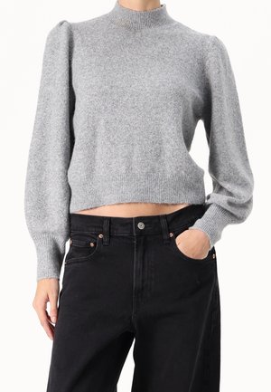 Personne portant un pull court gris clair à manches longues bouffantes et un jean noir taille haute, une main dans la poche.