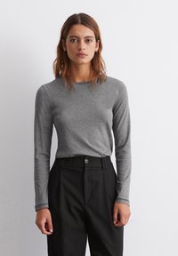 Cămașă gri cu mâneci lungi, realizată dintr-un material moale, având un guler rotund și un croi ajustat, purtată cu o pereche de pantaloni negri cu dungi fine.