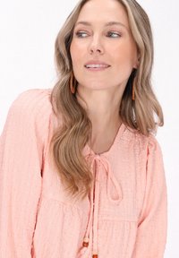  ferskenfarvet bluse med tekstur, lange ærmer, rund halsudskæring og bånd med perleindsatser. Bemærkelsesværdigt krøllemønster og afslappet pasform.