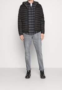 Doudoune noire avec capuche, dotée d'une fermeture éclair et d'une doublure verte. Portée sur une chemise à carreaux foncée, avec un jean gris clair retroussé et des bottes noires.