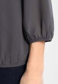 Graue Bluse mit Puffärmeln und elastischen Bündchen, strukturiertem Stoff und lässigem Schnitt am Saum, kombiniert mit dunkler Jeans.