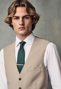 Gilet beige avec un col à revers sur une chemise blanche, complété par une cravate verte texturée et une barrette à cravate en argent, avec une seule poche et des boutons.
