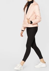 Jasnoróżowa kurtka puchowa na zamek z kapturem, pikowany wzór i srebrne akcenty, zestawiona z czarnymi legginsami i białymi trampkami.