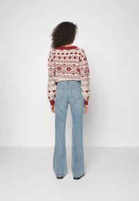 Pulôver tricotado vermelho e creme com padrões geométricos, combinado com jeans flare azul claro. Detalhes de costura visíveis e punhos ajustados.