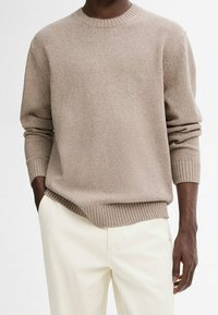 Homme portant un pull beige à col ras du cou et un pantalon blanc cassé, une main dans la poche, sur fond neutre.