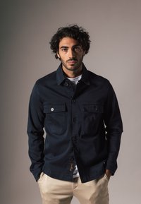 Donkerblauwe denimjas met knoop-geknopte borstzakken, een kraag en een rechte snit. Heeft een subtiele stikselafwerking aan de zoom.