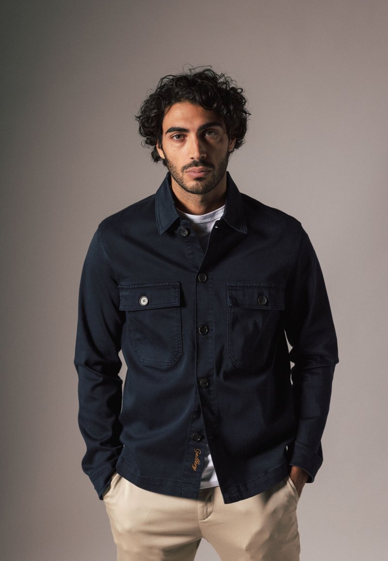 Donkerblauwe denimjas met knoop-geknopte borstzakken, een kraag en een rechte snit. Heeft een subtiele stikselafwerking aan de zoom.