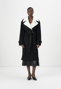 Manteau en laine noir avec un col blanc, taille ceinturée et couture contrastante. Présente une fermeture à un bouton et des poignets larges.