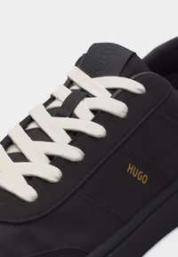 Baskets noires avec lacets blancs, logo "HUGO" doré sur le côté, et détail de boucle de lacet sur la languette.