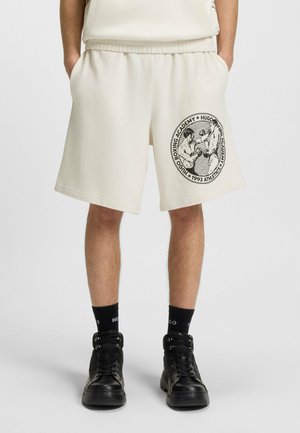 Man wearing cream shorts with a black boxing-themed graphic on the thigh, black socks, and black high-top sneakers, standing with hands in pockets.