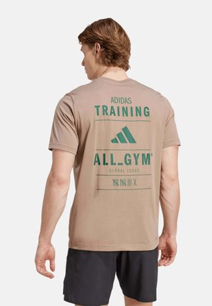 Joven con camiseta de entrenamiento beige de Adidas con logo y texto verde, combinada con pantalones cortos negros, de pie de espaldas.