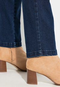 Zamszowe botki za kostkę w beżowym kolorze, z teksturowanym brązowym słupkowym obcasem. W połączeniu z ciemnoniebieskimi dżinsami, ukazującymi slim fit.