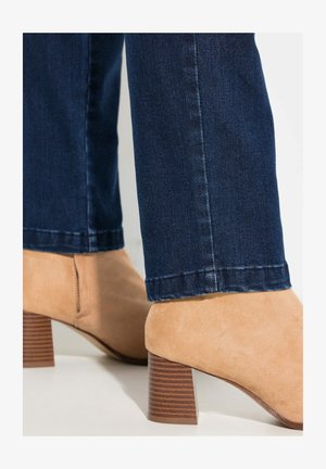 Beige Wildleder-Stiefelette mit einem strukturierten braunen Blockabsatz. Kombiniert mit dunkblauen Jeans im Slim-Fit.