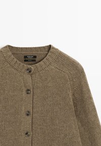 Cardigan en laine marron tricoté avec col rond et cinq boutons sombres sur le devant, montrant la partie supérieure du torse.