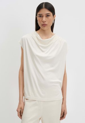 Femme aux cheveux raides et foncés portant un haut blanc drapé sans manches et un pantalon blanc, posant devant un fond gris clair.