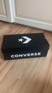 Fekete kartoncipős doboz fehér "CONVERSE" felirattal és logóval, csillaggal és nyíllal díszítve a fedélen, egy fa padlón áll.