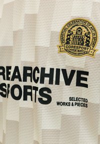 Tissu beige avec texte noir "REARCHIVE SPORTS SELECTED WORKS & PIECES" et un patch jaune portant l'inscription "Perpetual Celebration of Art in Sport COREPORT."