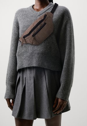 Persona que lleva un suéter gris texturizado, falda gris plisada y un bolso cruzado de pana marrón con dos cremalleras negras.