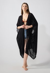 Musta läpikuultava kimono, jossa on leveät hihat, avoin etuosa ja kevyt kudotun laadun, käytetty ruskean bikinin ja farkkushortsien päällä.