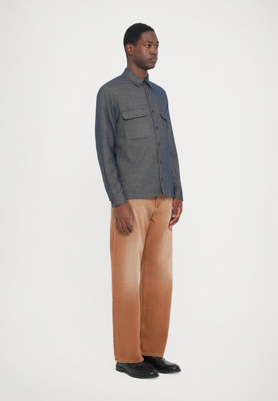 Cămașă gri cu nasturi, dotată cu două buzunare pe piept, asociată cu blugi largi din denim maro. Modelul poartă încălțăminte neagră. Design simplu și structurat.