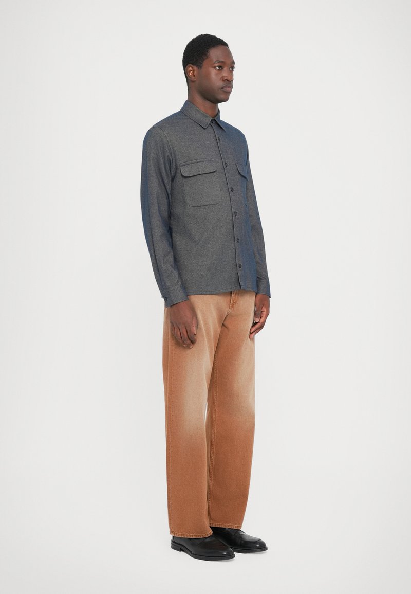 Camicia grigia con bottoni, dotata di due tasche sul petto, abbinata a jeans larghi marroni. Il modello indossa scarpe nere. Design semplice e strutturato.