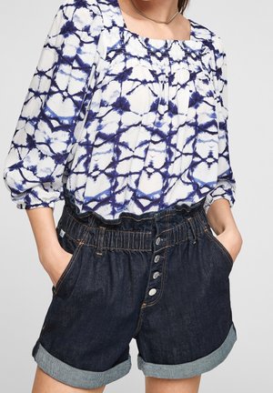 Hvid bluse med blå tie-dye mønster, firkantet udskæring og rynkede ærmer, kombineret med mørke denimshorts med rullet kant og knappedetalje.