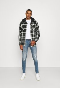 Camisa xadrez preto e branco com capuz preto, combinada com jeans rasgados azuis claros e sapatilhas brancas. T-shirt branca simples por baixo.