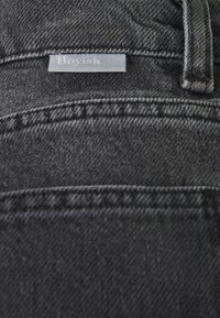 Tela de mezclilla negra con una textura sutil. Presenta una etiqueta que dice "Boyish", cosida a lo largo de la pretina, con un diseño de corte estándar.