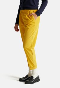 Gele corduroy broek met een relaxte pasvorm, elastische tailleband, zijzakken en opgerolde manchetten, gedragen met zwarte loafers en witte sokken.
