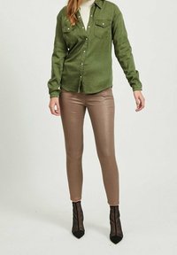 Oliwkowa koszula na guziki z dwoma kieszeniami na piersi, w połączeniu z dopasowanymi błyszczącymi brązowymi legginsami i przezroczystymi czarnymi botkami na obcasie.