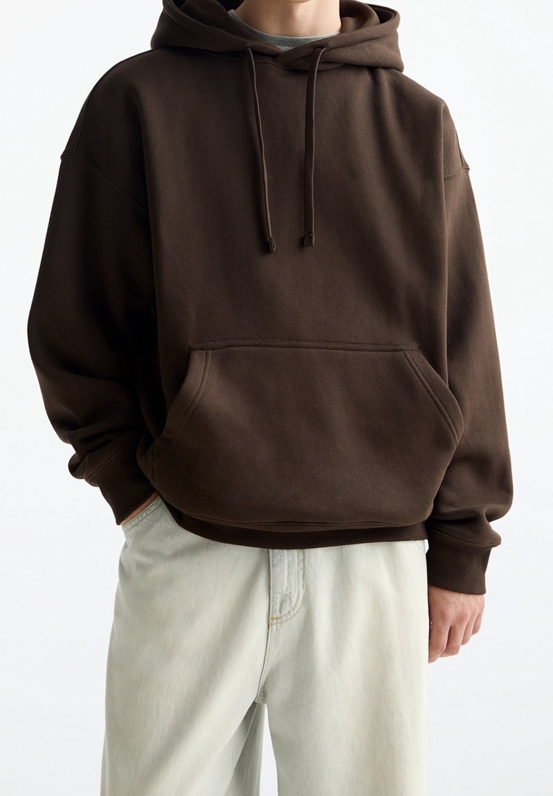 Felpa con cappuccio marrone, dotata di tasca frontale, vestibilità comoda, polsini e orlo a costine. Abbinata a un paio di pantaloni in denim chiaro.