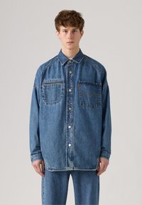 Løs-fittende denimskjorte i middels blå, med to brystlommer, knappelukking, og kontrasterende sømdetaljer. Teksturert stoff.
