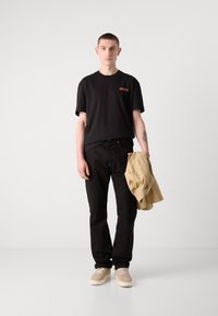 Musta puuvillainen t-paita, jossa on punainen logopatchi, mustat farkut ja vaaleanbeige kengät. Malli pitää vaaleanbeigeä takkia. Yksinkertainen, moderni muotoilu.
