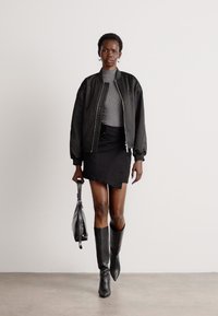 Giacca bomber nera, top grigio attillato, gonnellina di jeans nera, stivali neri fino al ginocchio, che porta una piccola borsa nera, sullo sfondo chiaro.
