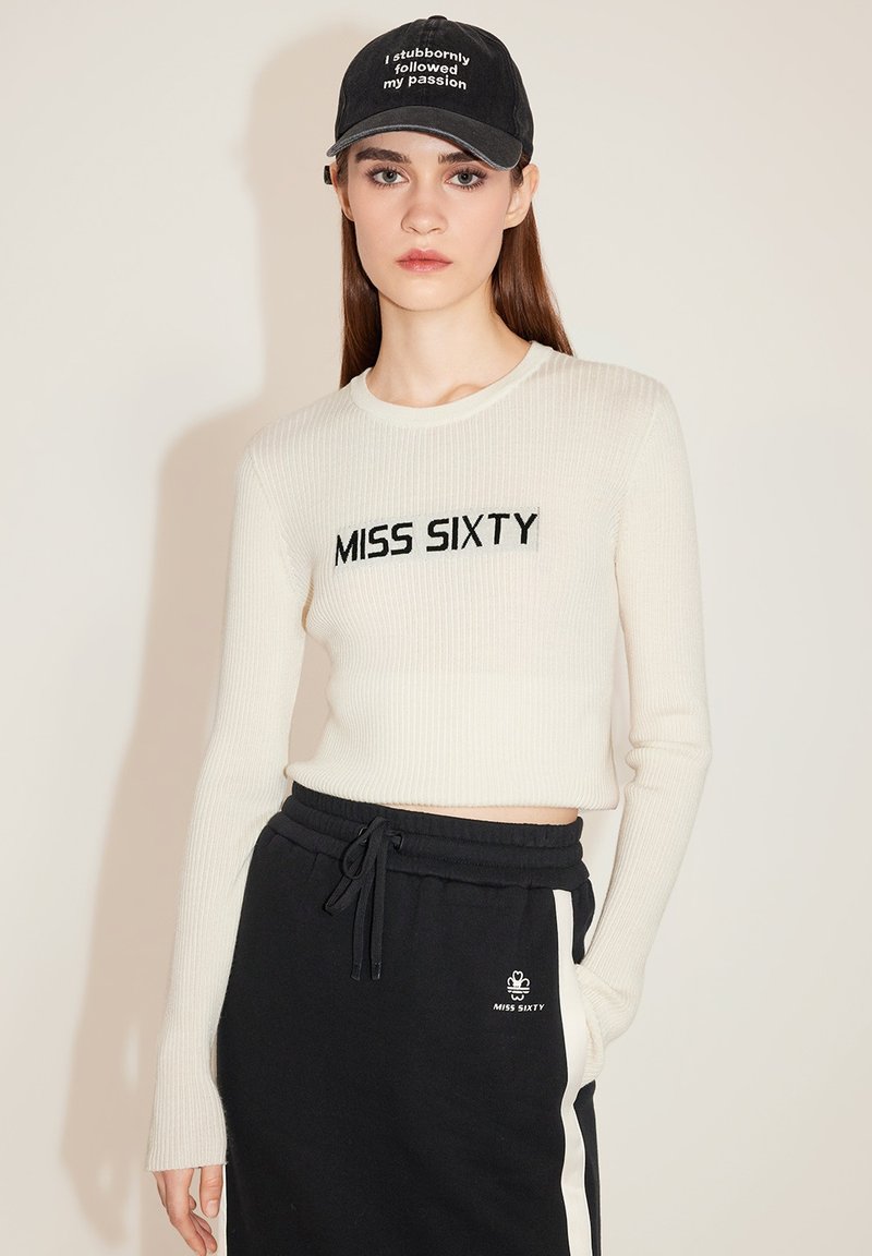 Cremfarbener, gerippter Langarm-Crop-Top mit schwarzem "MISS SIXTY"-Stick; kombiniert mit schwarzen Jogginghosen mit weißen Seitenstreifen und Logo.