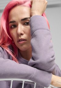 Sudadera de algodón morada con cuello redondo y puños acanalados. La modelo tiene el cabello lacio y rosa, y descansa su brazo en una silla blanca de alambre.