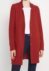 Rode wollen jas met een kraag, voorste drukknoopsluiting, zijzakken en lange mouwen, gedragen over een wit shirt en donkere spijkerbroek.