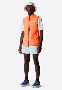 En orange lättviktsväst över en vit t-shirt med korta ärmar, i kombination med ljusblå shorts och blå sportskor. Västen har en svart dragkedja.