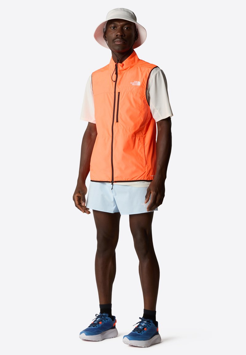 En orange lättviktsväst över en vit t-shirt med korta ärmar, i kombination med ljusblå shorts och blå sportskor. Västen har en svart dragkedja.