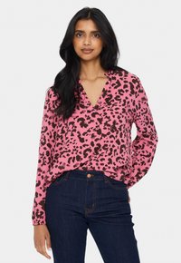 Kvinna med långt svart hår, iklädd en blus med rosa leopardmönster och mörkblå jeans, står med ena handen i fickan mot en vit bakgrund.