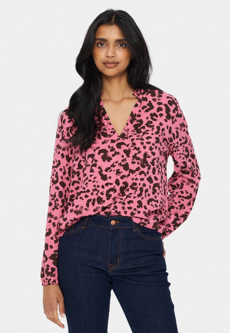 Kvinna med långt svart hår, iklädd en blus med rosa leopardmönster och mörkblå jeans, står med ena handen i fickan mot en vit bakgrund.