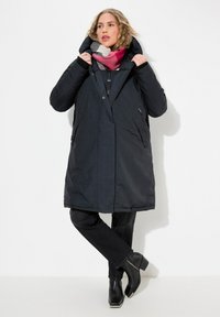 Abrigo largo negro con capucha, que cuenta con cierre de cremallera, dos bolsillos laterales y una textura suave. Combinado con una bufanda de rayas multicolores y pantalones negros.