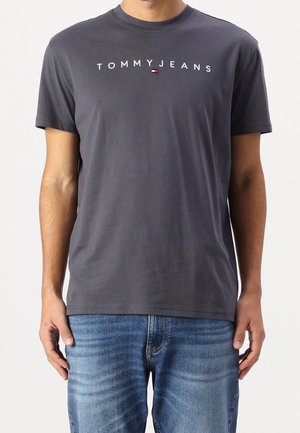 Man draagt een donkergrijs Tommy Jeans T-shirt gecombineerd met lichtblauwe versleten spijkerbroek, staand met armen ontspannen langs het lichaam.