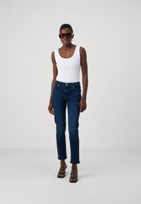 Débardeur blanc à côtes, jean skinny bleu foncé, sandales à brides noires, lunettes de soleil surdimensionnées de couleur sombre, texture lisse et coupe ajustée.