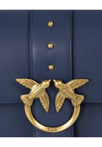 Navy modra usnjena torbica s poudarkom iz zlatega obroča z dvema pticama. Okrasne zlate žebljičke krasijo osrednji trak. Površina je gladka.