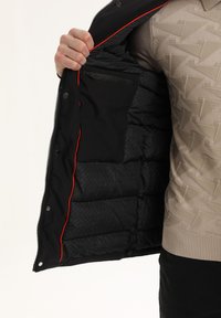 Giacca a vento nera con interni rossi, dotata di tasca con zip e texture trapuntata. L'esterno è liscio e resistente all'acqua.